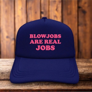 May include: A navy blue baseball cap featuring the text BLOWJOBS ARE REAL JOBS printed in bold pink capital letters across the front panel. The cap has a structured crown and a curved brim, maintaining a consistent color scheme throughout the design.