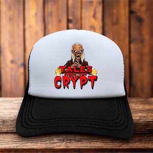 Może przedstawiać: Biało-czarna czapka typu trucker z grafiką postaci Crypt Keeper z Tales from the Crypt. Projekt zawiera czerwony napis "TALES FROM THE CRYPT" z efektem ściekającej czcionki oraz dwie żółte świece po obu stronach.