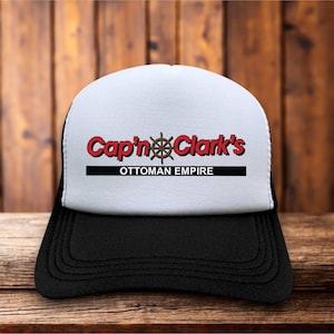 Puede incluir: Una gorra tipo camionero con panel frontal blanco y visera negra. El frente muestra el texto rojo Cap'n Clark's con un icono de timón de barco marrón reemplazando el apóstrofo, una línea negra y el texto OTTOMAN EMPIRE en letras negras.