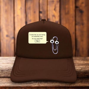 May include: A brown baseball cap featuring a graphic of a silver paperclip character with large eyes. Next to the character is a pale yellow speech bubble containing the text: It looks like you are having an existential crisis. Do you need help? Yes.