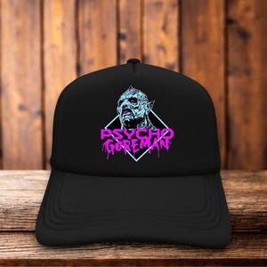 May include: A black baseball cap featuring a graphic print of a creature's head in light blue and purple, centered above the text "PSYCHO GOREMAN" in bright pink, all enclosed within a light blue diamond outline on the front of the cap.