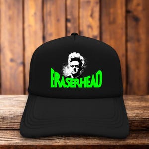 May include: A black baseball cap featuring a white graphic of a person with spiky hair above the bright green text Eraserhead. The cap has a structured front panel and a curved brim, displaying a bold contrast between the black fabric and the design.
