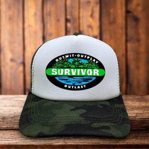 May include: A white foam front trucker hat with a green camouflage brim and mesh back. The front features a black oval logo with the text SURVIVOR in white, surrounded by green palm trees, blue water graphics, and the words OUTWIT OUTPLAY OUTLAST.