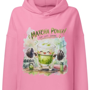 Op de afbeelding: Een felroze hoodie met een aquarel-stijl afbeelding van een glimlachende matcha-drank die een halter optilt. De tekst boven de illustratie luidt "MATCHA POWER! SIP. LIFT. SHINE.". Het ontwerp bevat ook een kettlebell en een shakerbeker.