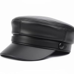 Op de afbeelding: Zwarte leren kapiteinshoed met een korte klep en een decoratieve band. De hoed heeft een knoopdetail aan de zijkant en staat tegen een witte achtergrond. Een klassieke hoedenstijl.