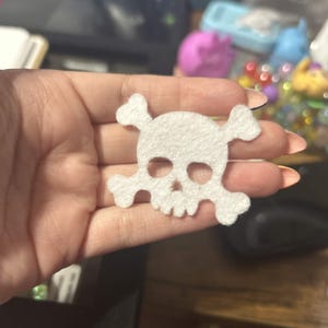 May include: A white felt skull and crossbones applique with cut-out eye sockets and a nasal cavity. The skull features a rounded cranium and a jagged jawline, while the crossbones extend from behind the skull on both the left and right sides.