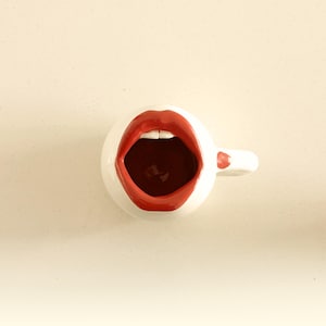 May include: A white ceramic mug with a red lip design. The open mouth design features red lips and white teeth. The mug has a small handle and is set against a neutral background.