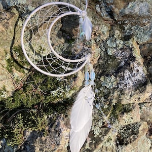 May include: A white crescent moon dreamcatcher with a woven web design. The dreamcatcher features white feathers, clear beads, and a string of iridescent beads. The background is a natural stone surface with moss.