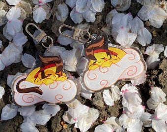 May include: Two acrylic keychains featuring a cartoon character sleeping on a cloud. The character has yellow skin, brown hair, and a red headband. The keychains have silver-colored clasps and are surrounded by white flower petals.