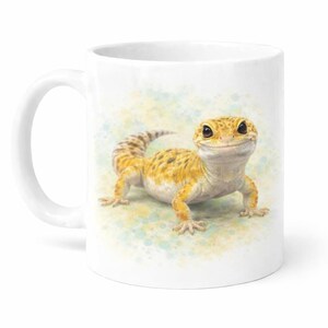 May include: White ceramic mug with a detailed illustration of a smiling yellow and brown leopard gecko. The gecko is centered on the mug, with a watercolour-style background of blue and yellow spots. The mug has a curved handle.