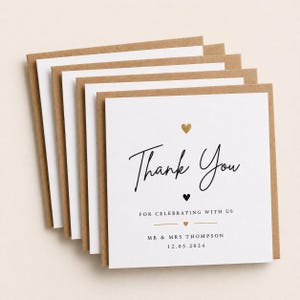 May include: A stack of thank you cards with a white interior and a brown kraft paper border. The cards feature the words "Thank You" in elegant script, with a gold heart and a black heart. The text "For celebrating with us" is also included.