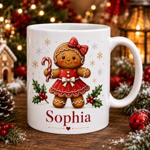 Könnte beinhalten: Ein weißer Keramikbecher mit einer Lebkuchenfigur-Illustration, die ein rotes Kleid und eine rote Schleife trägt und eine Zuckerstange hält. Das Design zeigt Stechpalmenblätter, Beeren und Schneeflocken. Der Name Sophia steht in rotem Text darunter.