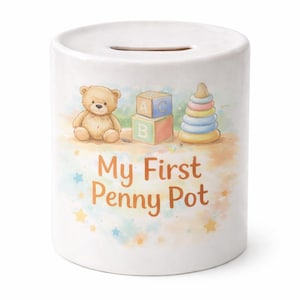Puede incluir: Hucha de cerámica blanca con una ranura en la parte superior. Está decorada con un osito de peluche, bloques de construcción y un juguete apilable. El texto "My First Penny Pot" está impreso en la parte delantera, con estrellas de colores y detalles de acuarela.