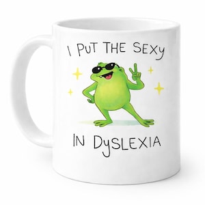 May include: White ceramic mug with a cartoon frog wearing sunglasses and a peace sign. The text on the mug reads "I PUT THE SEXY IN DYSLEXIA". The design also includes yellow star accents.