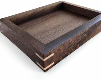May include: A rectangular wooden tray with a dark brown finish. The tray has a raised border and lighter-colored accents at the corners. The wood grain is visible, adding texture to the piece. This tray is suitable for home decor.
