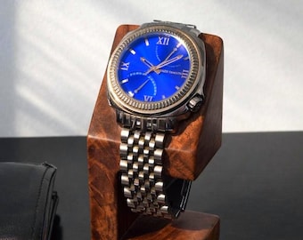 May include: A silver wristwatch with a blue face and Roman numeral hour markers is displayed on a wooden watch stand. The watch has a silver band and a gold-colored bezel. The stand is made of dark wood.