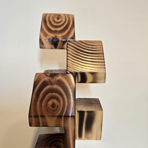 May include: A wooden sculpture composed of stacked, geometric blocks. The blocks are various sizes and shapes, with visible wood grain and a burnt finish. The sculpture is a warm brown color, with dark charring on the edges, creating a contrast.