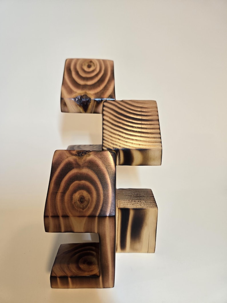 May include: A wooden sculpture composed of stacked, geometric blocks. The blocks are various sizes and shapes, with visible wood grain and a burnt finish. The sculpture is a warm brown color, with dark charring on the edges, creating a contrast.