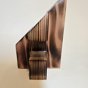 May include: A modern, abstract wooden sculpture with a geometric design. The sculpture features a triangular shape and a rectangular base, with vertical lines carved into the wood. The wood has a warm, brown tone.
