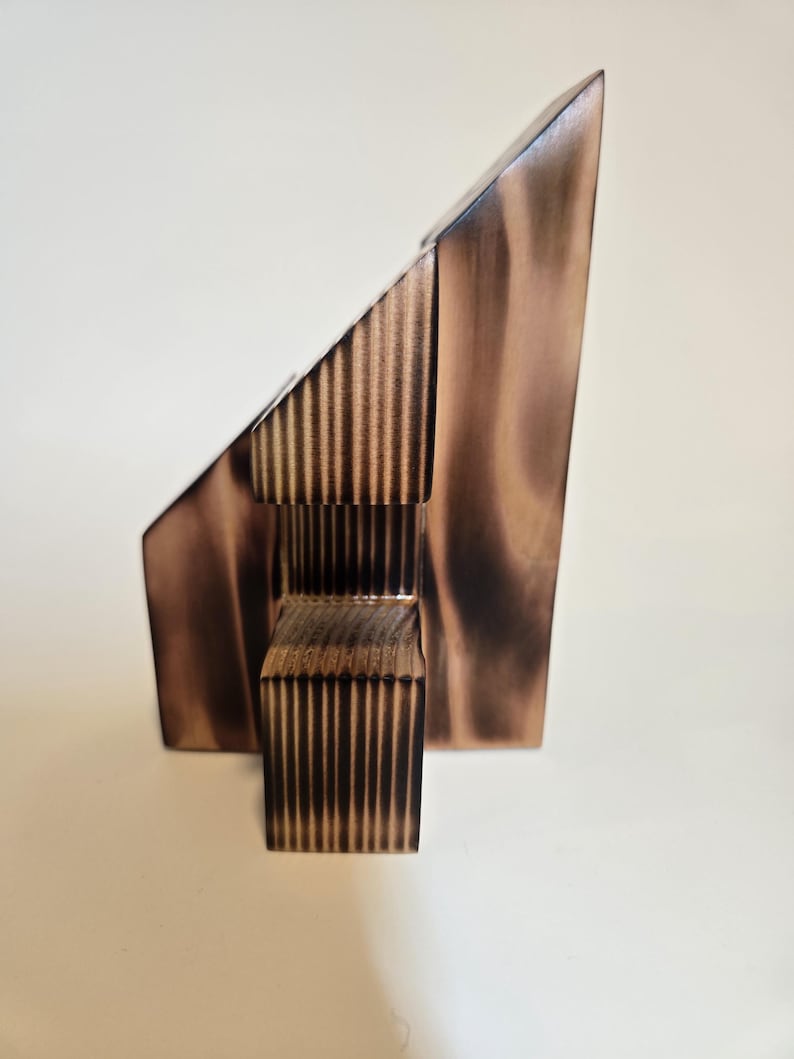 May include: A modern, abstract wooden sculpture with a geometric design. The sculpture features a triangular shape and a rectangular base, with vertical lines carved into the wood. The wood has a warm, brown tone.