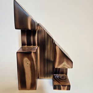 May include: Abstract wooden sculpture with geometric shapes. The sculpture features a combination of rectangular, triangular, and vertical elements, with a dark brown finish and visible wood grain.