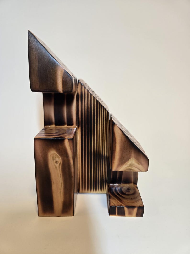 May include: Abstract wooden sculpture with geometric shapes. The sculpture features a combination of rectangular, triangular, and vertical elements, with a dark brown finish and visible wood grain.