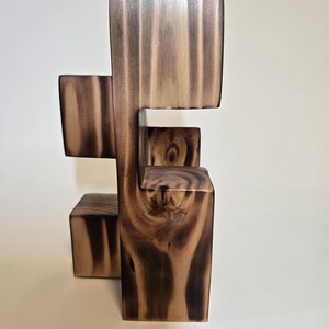 May include: A modern wooden sculpture with a unique, geometric design. The sculpture is constructed from blocks of wood, showcasing a natural wood grain pattern in shades of brown and tan. The piece has a minimalist aesthetic.