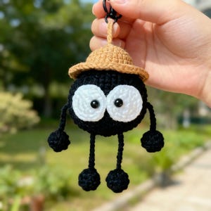 Może przedstawiać: Ręcznie robiony brelok do kluczy z szydełka przedstawiający czarną postać z dużymi białymi oczami i małym brązowym kapeluszem. Brelok ma zwisające ramiona i nogi z czarnymi pomponami. Dołączony jest czarny metalowy zatrzask.
