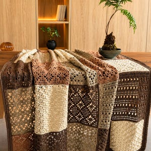 Può includere: Una coperta all'uncinetto fatta a mano nei toni del marrone, beige e marrone chiaro. La coperta presenta un design patchwork con vari motivi traforati, tra cui motivi floreali e geometrici. La coperta è drappeggiata su una superficie di legno.