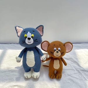 May include: A blue and white crocheted cat doll with yellow eyes and pink inner ears standing next to a brown and tan crocheted mouse doll with large pink inner ears and black whiskers. Both handmade figures feature stitched facial details.