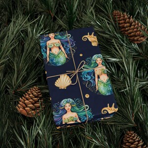 May include: Dark blue wrapping paper featuring illustrations of mermaids with blue and green hair, seashell accents, and gold dots. The paper is tied with twine and set against a backdrop of pine needles and pine cones.