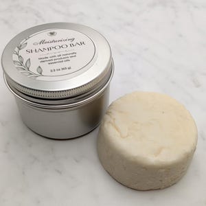 May include: A silver tin with a lid, next to a round, off-white shampoo bar. The tin's label reads "Moisturizing SHAMPOO BAR" and "2.2 oz (63 g)". The label also states the bar is made with naturally derived products and essential oils.