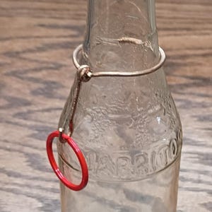 May include: Clear glass bottle with a wire closure and a red ring. The bottle has embossed lettering and is sitting on a wooden surface. The bottle is empty.