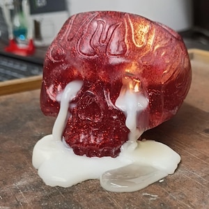 Puede incluir: Una vela de calavera roja con purpurina y detalles de llama en la parte superior. La vela está encendida, con cera blanca goteando de las cuencas de los ojos y acumulándose en la base. La calavera está sobre una superficie de madera.