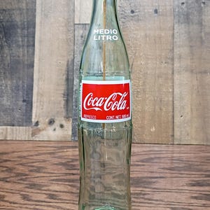 May include: A clear green glass Coca-Cola bottle with a red and white label reading Medio Litro and Cont. Net. 500 ml. A thin yellow incense stick stands upright inside the bottle, topped with a small red metal ring at the bottle opening.