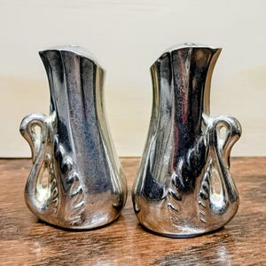 May include: Two silver-colored salt and pepper shakers. Each shaker has a curved handle and a decorative leaf pattern. The shakers are on a wooden surface.