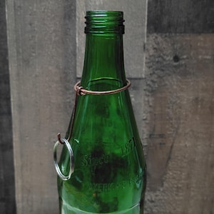 Puede incluir: Una botella de vidrio verde con cierre de alambre de cobre y un anillo de metal plateado. La botella tiene el texto "Since 1871 AMERICAN PREMIUM" en relieve en la parte delantera. La botella está vacía y tiene una estética vintage.