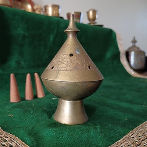 May include: A brass incense burner with a conical lid featuring etched floral patterns and small circular ventilation holes. The burner has a rounded body and a flared pedestal base. Three small brown incense cones sit to the left.