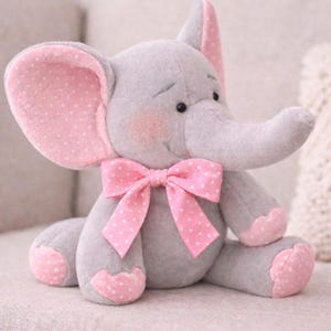 Könnte beinhalten: Ein graues Plüschelefant mit rosa gepunkteten Ohren, Füßen und einer Schleife. Das Stofftier ist 24 cm groß und trägt den Text "PDF PATTERN + Tutorial".