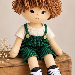 Puede incluir: Una muñeca de tela con cabello de lana marrón, rasgos faciales bordados y una camiseta color crema. La muñeca viste un overol de cuadros verdes y negros con dos botones verdes pequeños en los tirantes y zapatillas negras con cordones blancos.