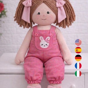 Peut inclure: Une poupée faite à la main, de 55 cm de haut, avec des cheveux bruns en fil de laine coiffés en couettes ornées de nœuds roses. La poupée porte une salopette rose avec un appliqué de lapin et des chaussures assorties à nœuds. La poupée a un visage souriant.
