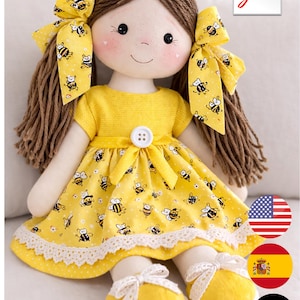 Op de afbeelding: Een handgemaakte pop met bruin garen haar en een gele jurk versierd met bijenpatronen. De pop, met een afmeting van 43 cm (17 inch), heeft een lief gezicht met roze wangen en draagt gele schoenen met witte strikken. De jurk heeft een witte kanten rand.