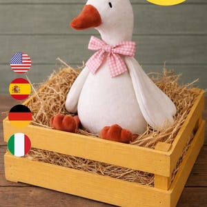 Può includere: Un'oca di peluche bianca con un papillon rosa a quadretti e becco e zampe arancioni, adagiata in una cassa di legno gialla piena di paglia. L'immagine include il testo "PDF Pattern + Tutorial" e "3 Sizes Goose".