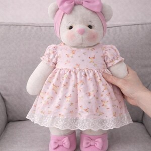 May include: A plush white teddy bear wearing a pink dress with a floral pattern, a matching headband with a bow, and pink shoes with bows. The dress has a white lace trim. The bear has rosy cheeks and a pink nose.