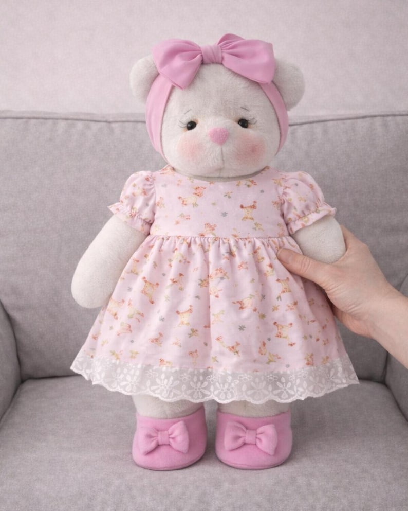 May include: A plush white teddy bear wearing a pink dress with a floral pattern, a matching headband with a bow, and pink shoes with bows. The dress has a white lace trim. The bear has rosy cheeks and a pink nose.
