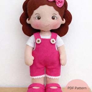 May include: A handmade doll with brown hair and rosy cheeks, wearing a white shirt and pink overalls with daisy accents. The doll has a pink bow and matching shoes. The doll is standing on a wooden surface.