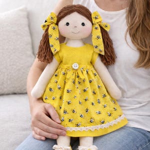 May include: A handmade cloth doll with brown yarn hair styled in pigtails, adorned with yellow bows. The doll wears a yellow dress patterned with bees, a white lace trim, and yellow shoes with white bows. The doll is being held.