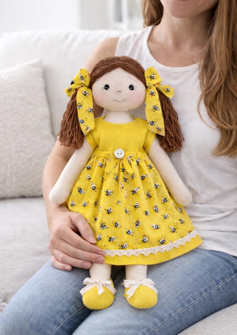 May include: A handmade cloth doll with brown yarn hair styled in pigtails, adorned with yellow bows. The doll wears a yellow dress patterned with bees, a white lace trim, and yellow shoes with white bows. The doll is being held.