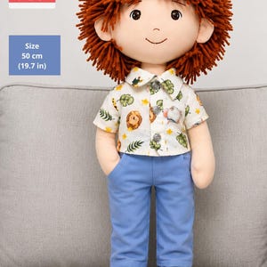 May include: A fabric doll with textured rust-colored yarn hair, wearing a white short-sleeved button-down shirt with a colorful animal and leaf print, blue trousers, and black and white sneakers. The doll measures 19.7 inches in height.