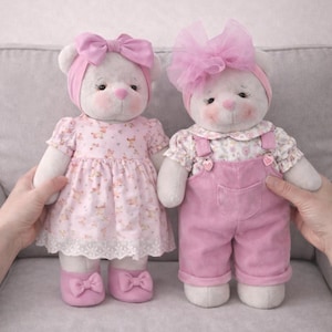 May include: Two plush teddy bears, each with white fur, rosy cheeks, and pink accents. One wears a pink dress and shoes with bows, the other wears pink overalls. Both have pink headbands and bows.